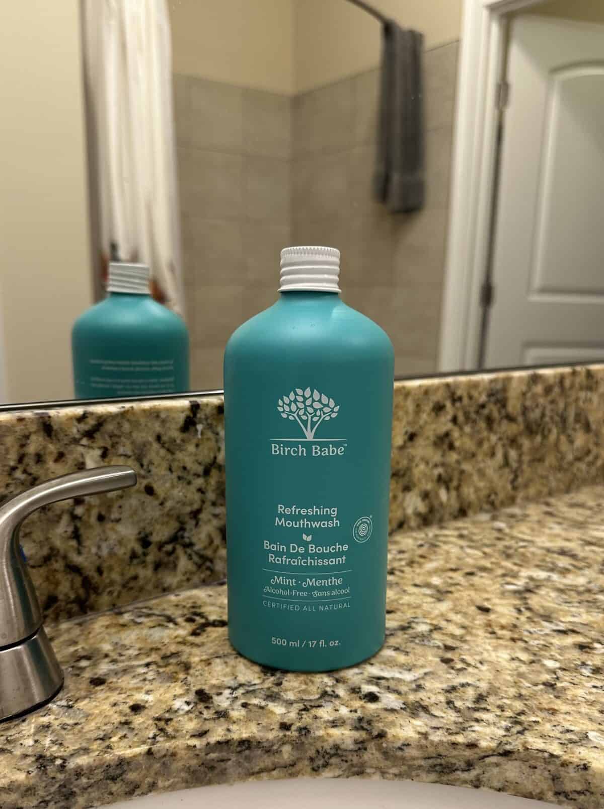 The photo shows a large teal bottle of Birch Babe "Refreshing Mouthwash" sitting on a speckled granite bathroom countertop in front of a mirror.