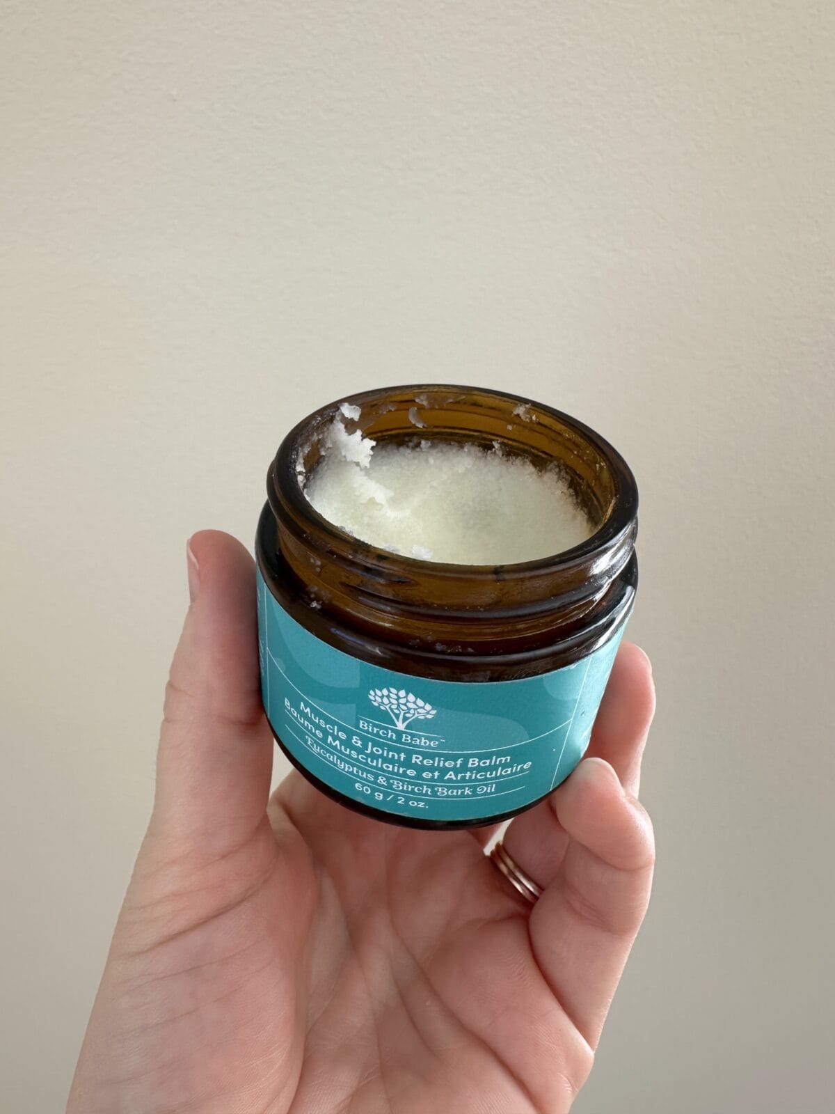 The photo shows a hand holding an open amber glass jar of Birch Babe "Muscle & Joint Relief Balm" with eucalyptus and birch bark oil, revealing the textured white balm inside against a plain wall.