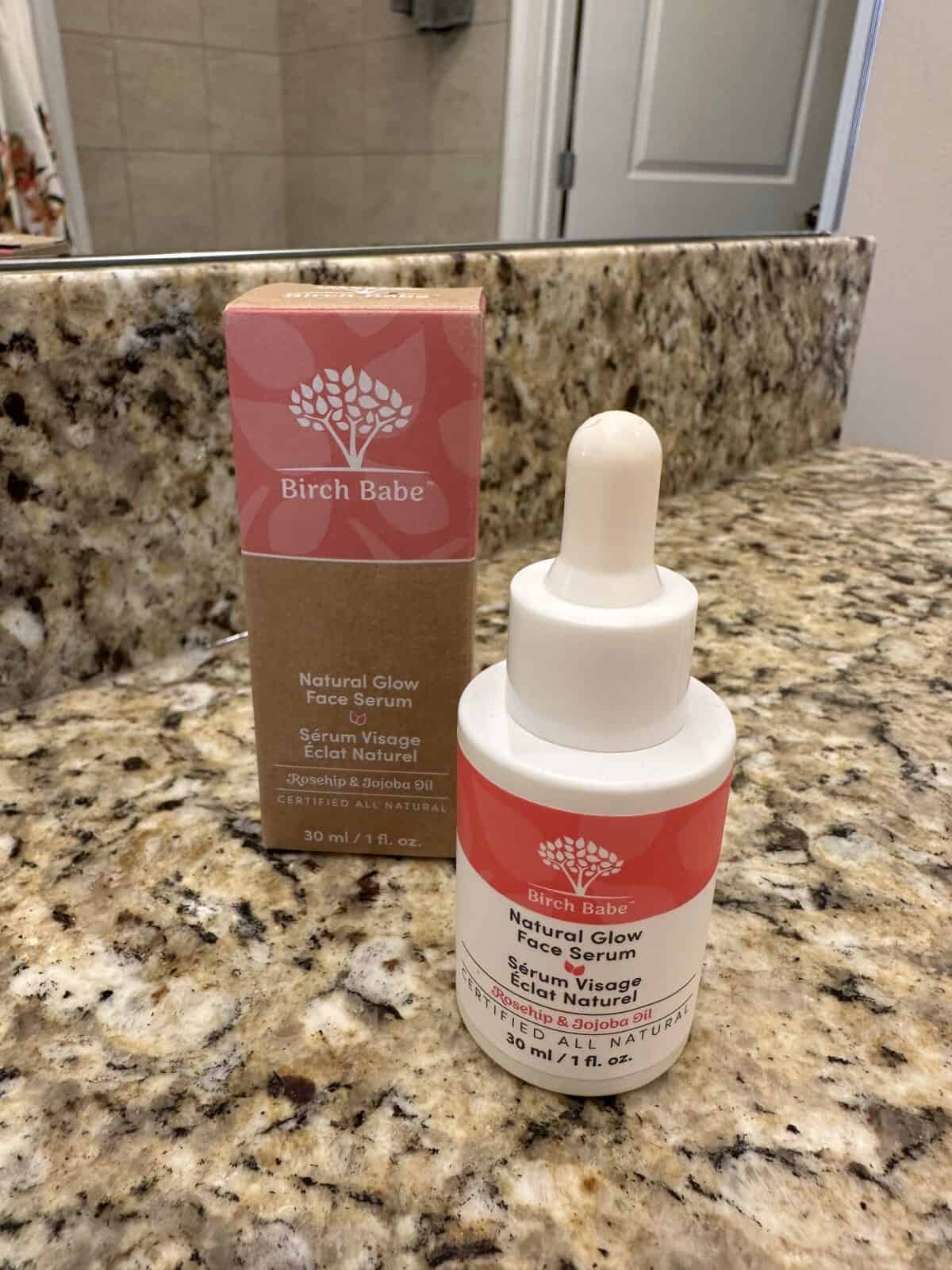 The photo shows a bottle and box of Birch Babe "Natural Glow Face Serum" sitting on a speckled bathroom countertop in front of a mirror.