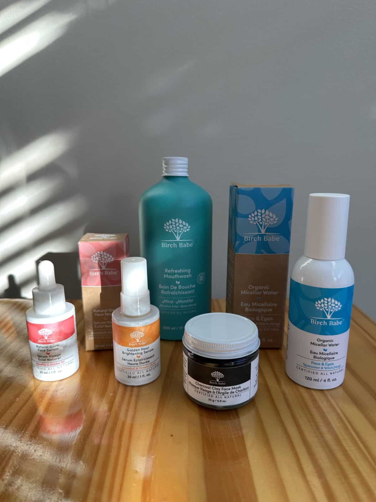 The photo displays an assortment of Birch Babe natural skincare and hygiene products, including serums, mouthwash, a charcoal face mask, and micellar water, arranged on a light wooden surface.