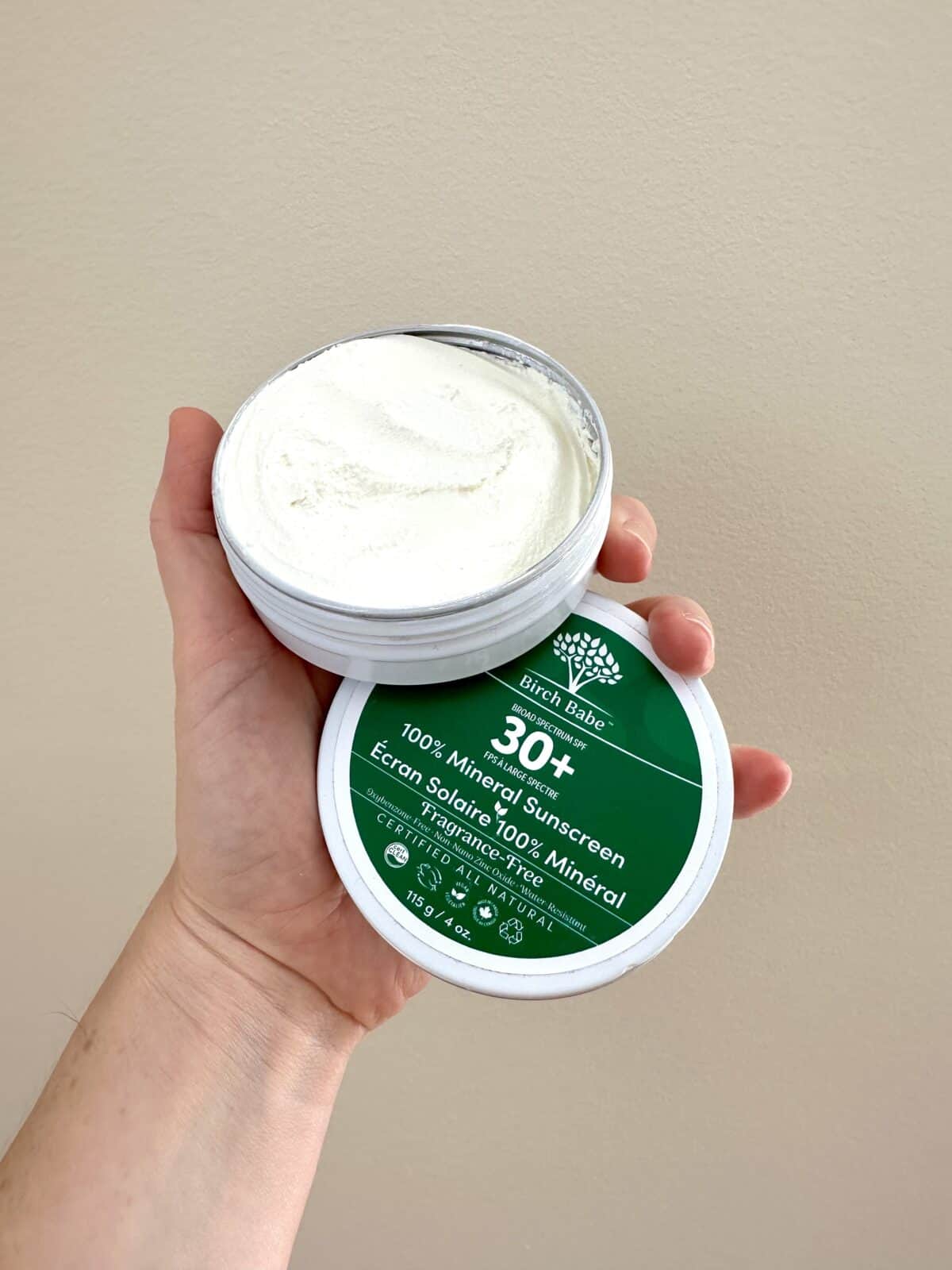 The photo shows a hand holding an open tin of Birch Babe "100% Mineral Sunscreen" with SPF 30+, revealing the thick white cream inside against a plain, neutral-colored wall.