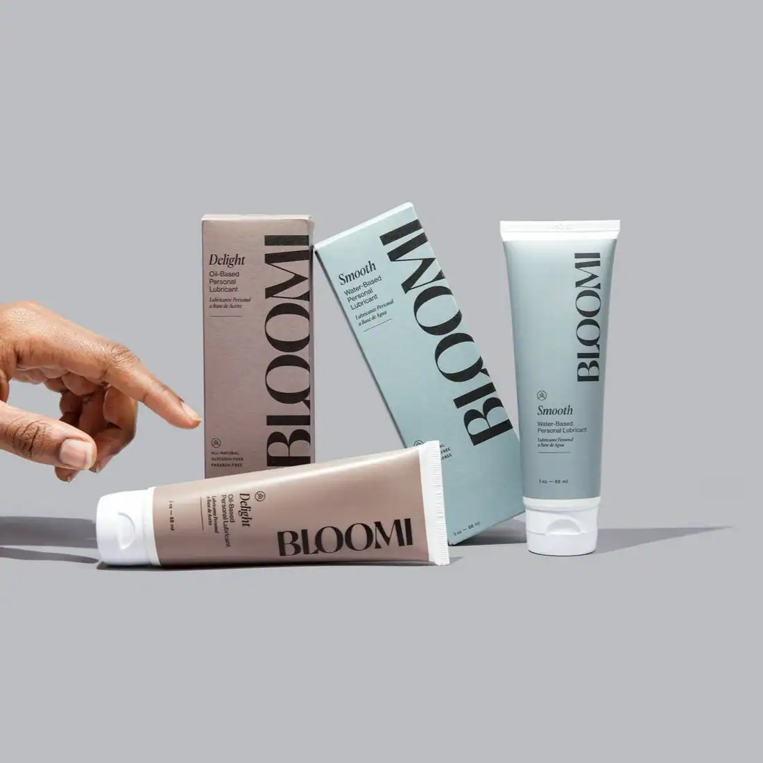 A still-life photo displays two types of Bloomi personal lubricants, "Delight" (oil-based) and "Smooth" (water-based), with their tubes and boxes arranged on a gray background, and a finger pointing toward the products.