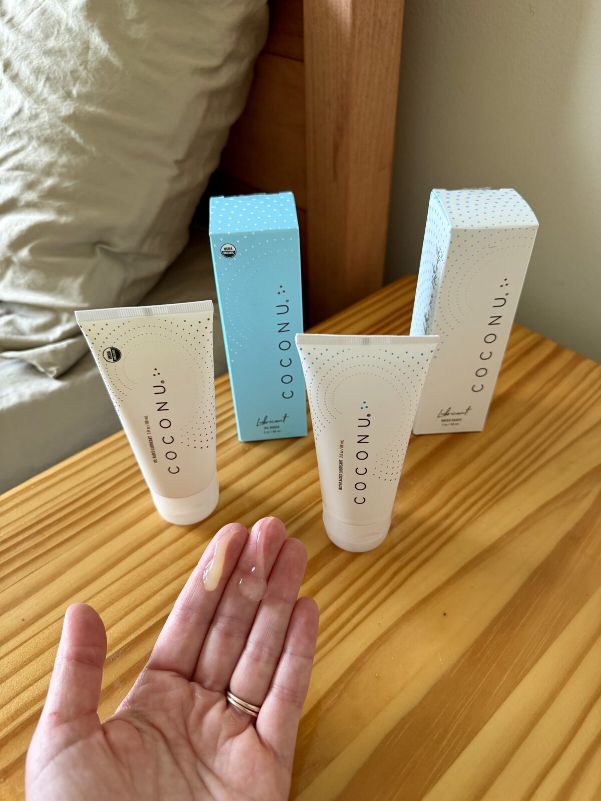 A hand with a small amount of cream faces up in front of two tubes labeled COCONU and their boxes on a wooden nightstand beside a bed.