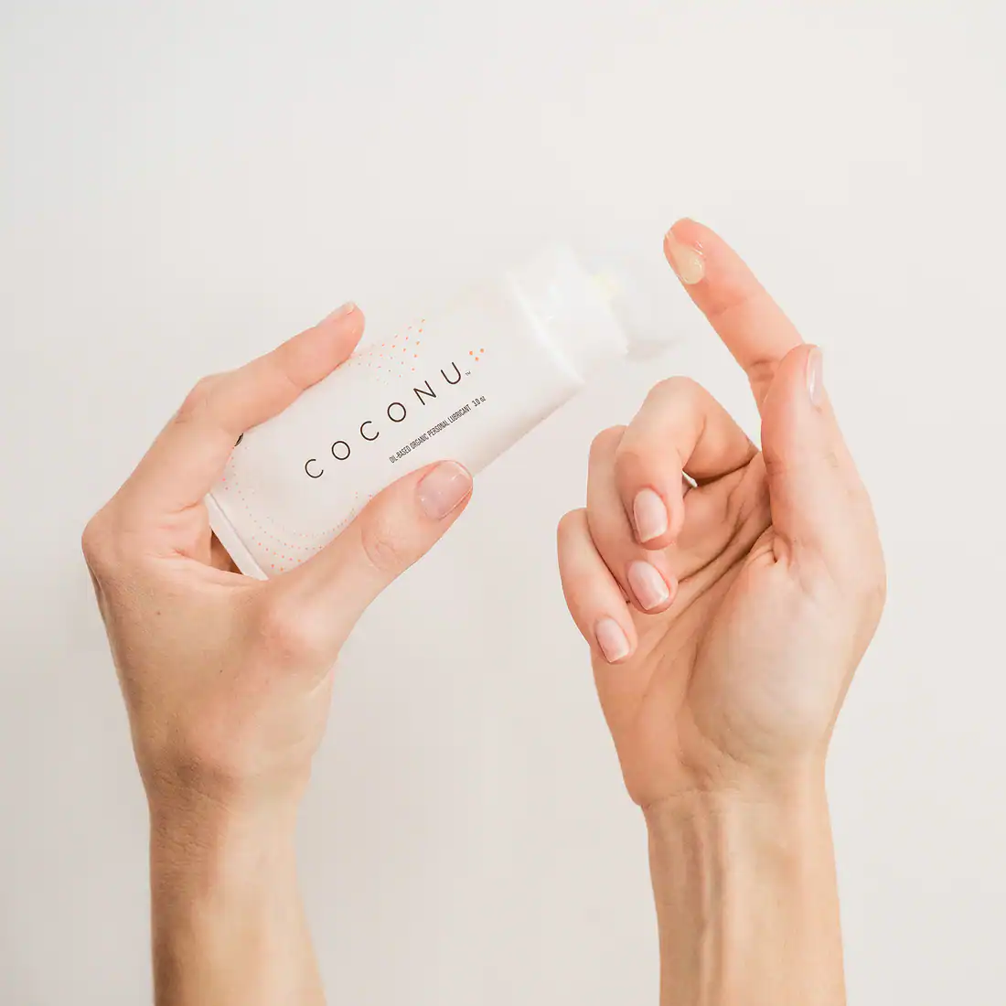 A hand is holding a white tube of COCONU oil-based organic personal lubricant, with a small amount of the product dabbed on the tip of an index finger.