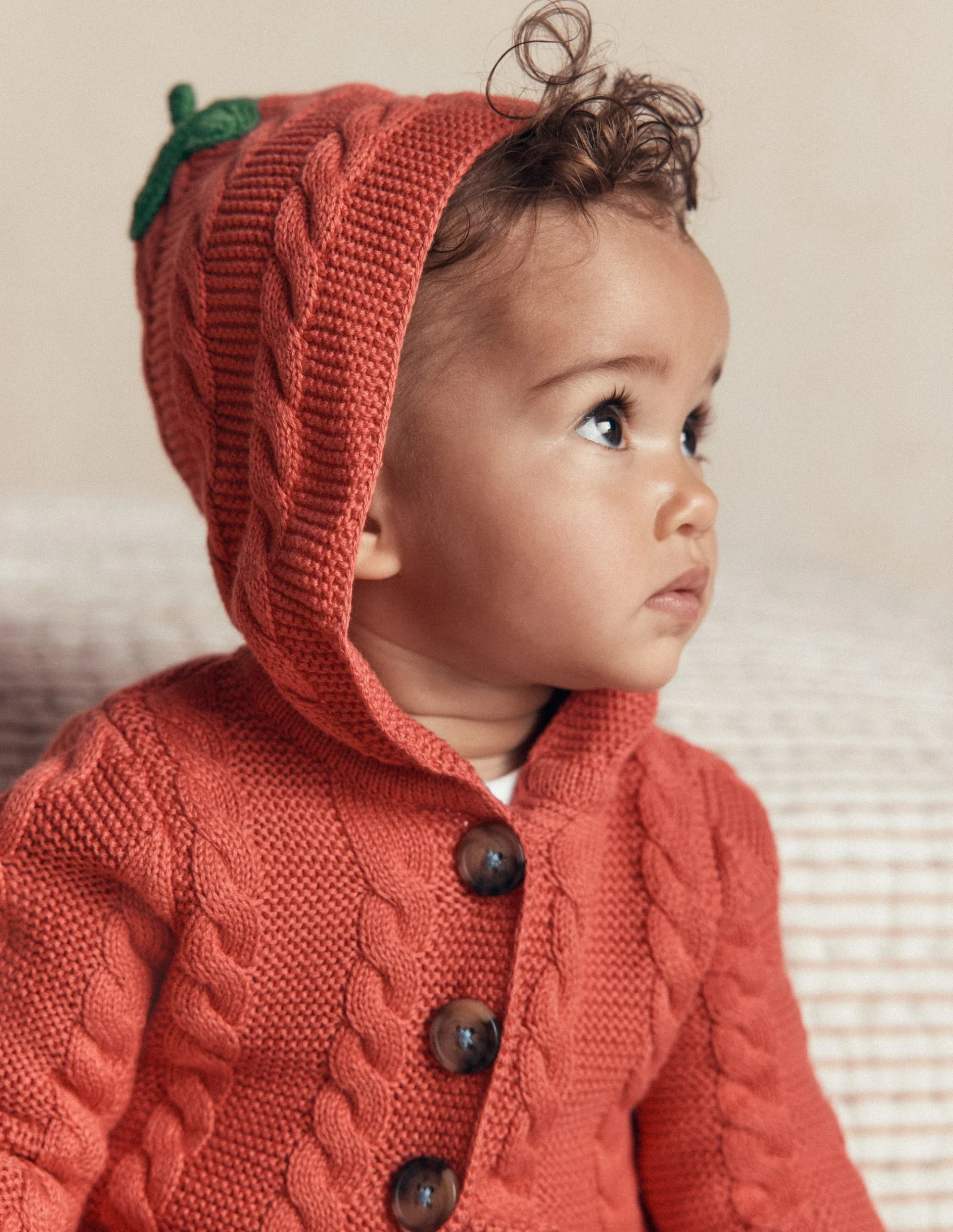 A toddler wearing a knitted orange hooded sweater with a green stem detail looks up and to the side. The sweater features large brown buttons and a cable-knit pattern. The child is sitting on a light-colored textured surface.