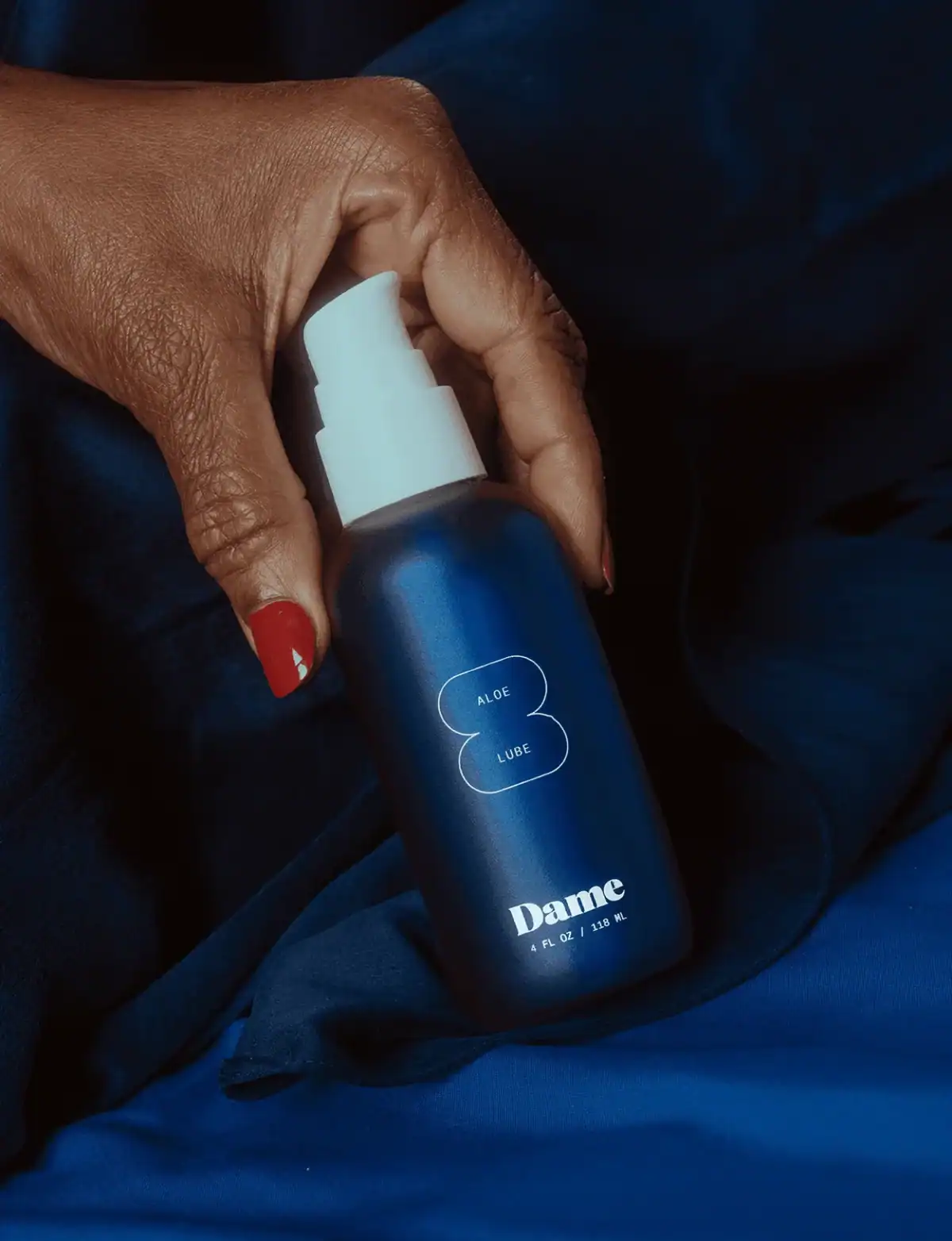 A close-up of a dark-skinned hand with red nail polish is holding a sleek, dark blue pump bottle of Dame Aloe Lube against a rippling, deep blue satin fabric background.