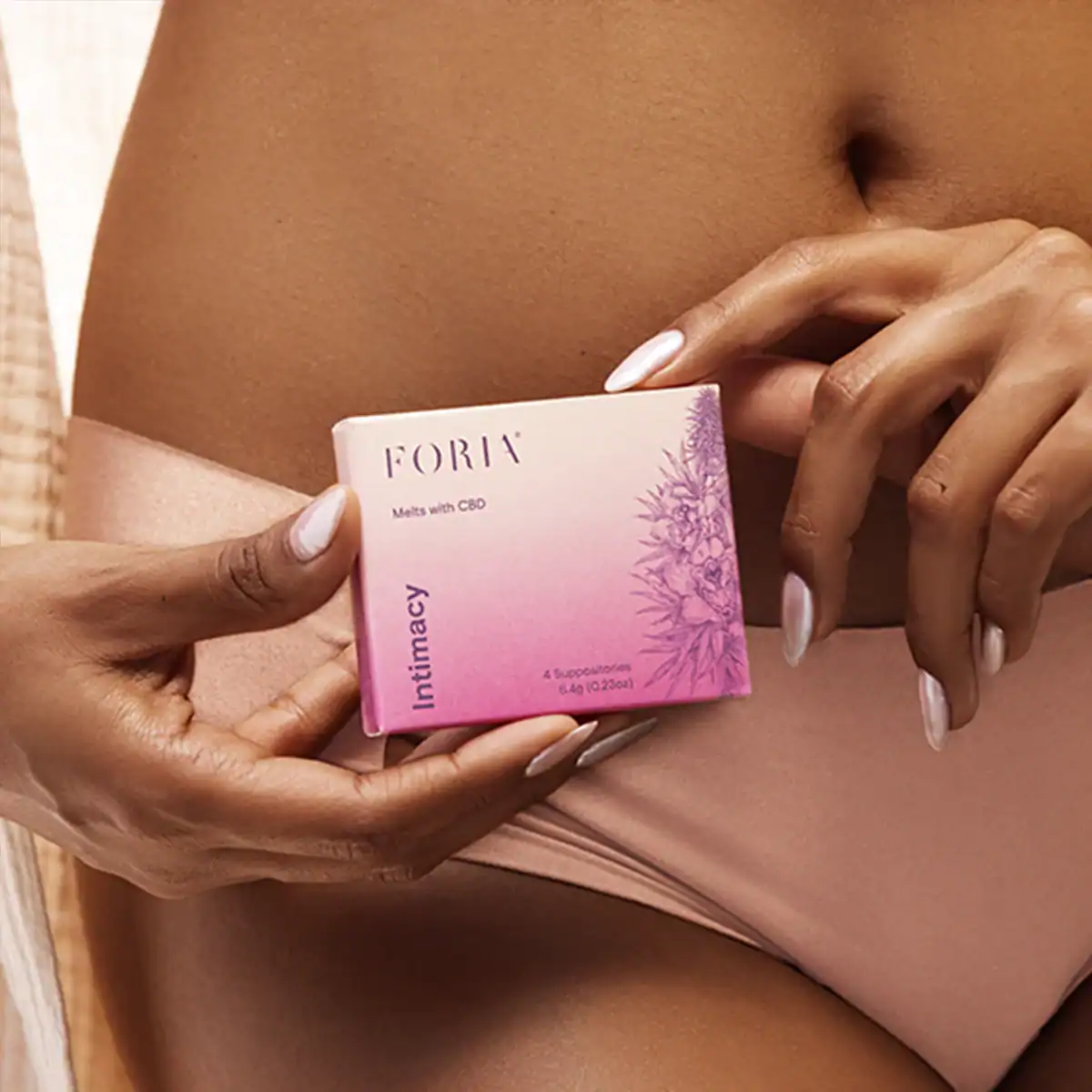 A person is holding a small, pink and white box of Foria Intimacy Melts with CBD in their hands, placed against their stomach area above light pink underwear.