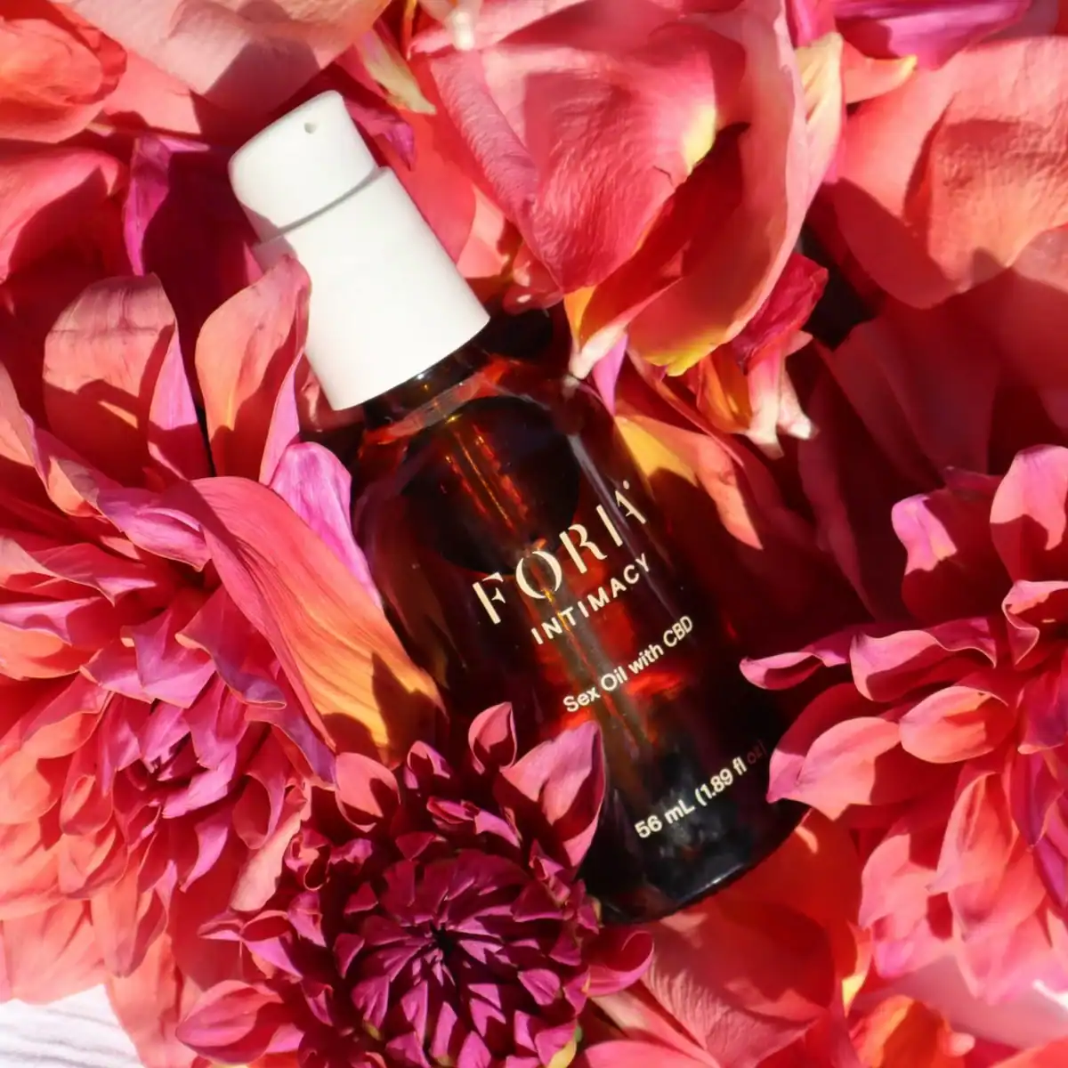 A bottle of Foria Intimacy Sex Oil with CBD is nestled on a bed of bright pink and reddish dahlia flower petals.
