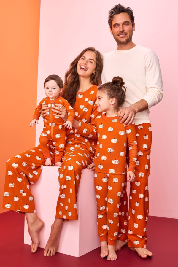 A family of four poses together. The adults, a baby, and a young child all wear matching orange pajamas with white pumpkin patterns, except the man, who wears matching pants and a white shirt. The background is pink and peach.