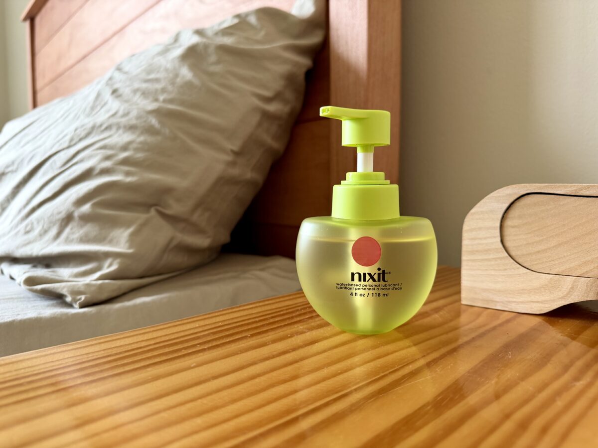 A nixit foaming wash bottle with a pump sits on a wooden nightstand next to a bed with beige sheets and a wooden headboard. A clock is also partially visible on the nightstand.