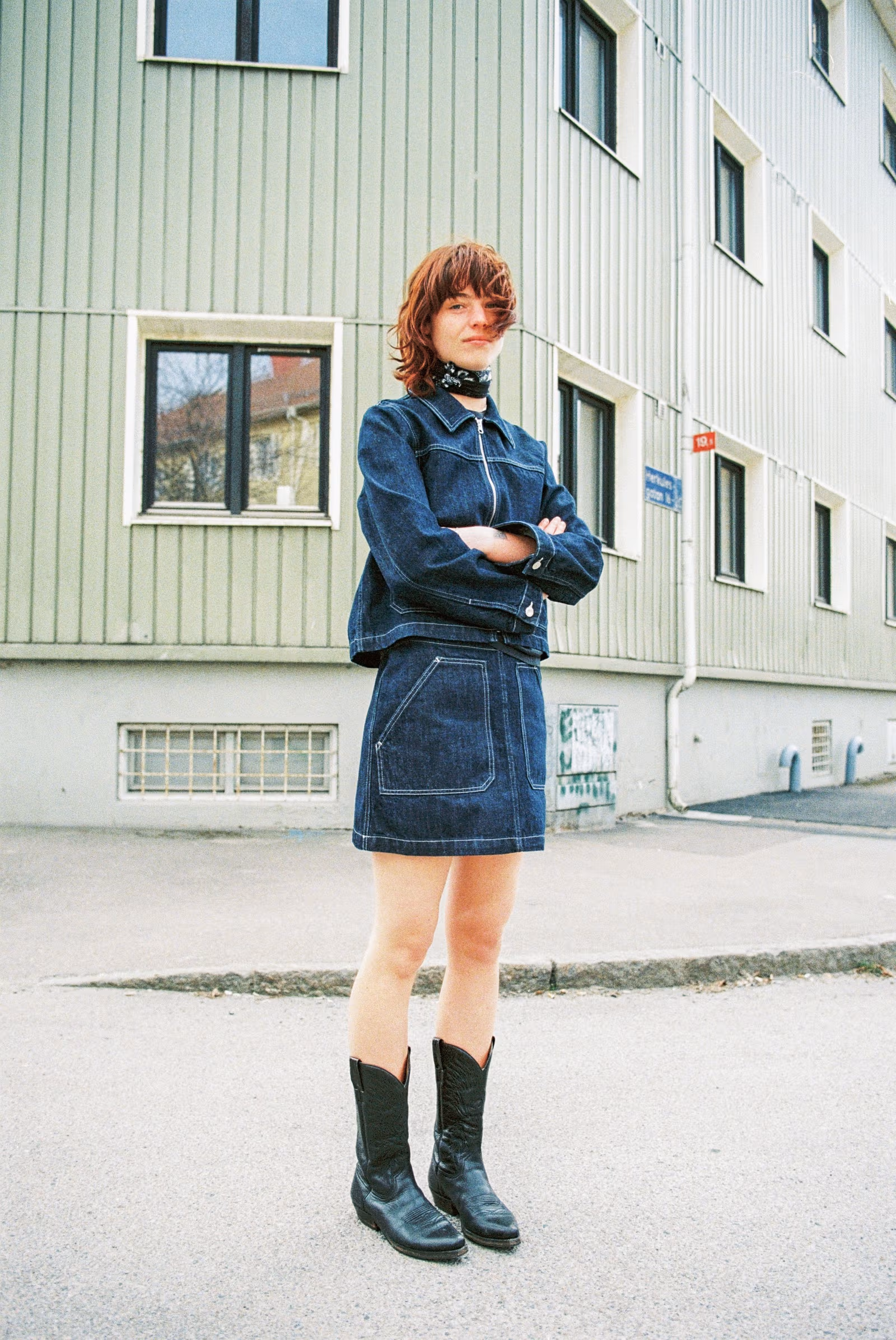 A person with short reddish hair and a scarf is standing on a street with their arms crossed, wearing a dark blue denim jacket from Nudie Jeans, a matching denim skirt, and black cowboy boots, in front of an apartment building.