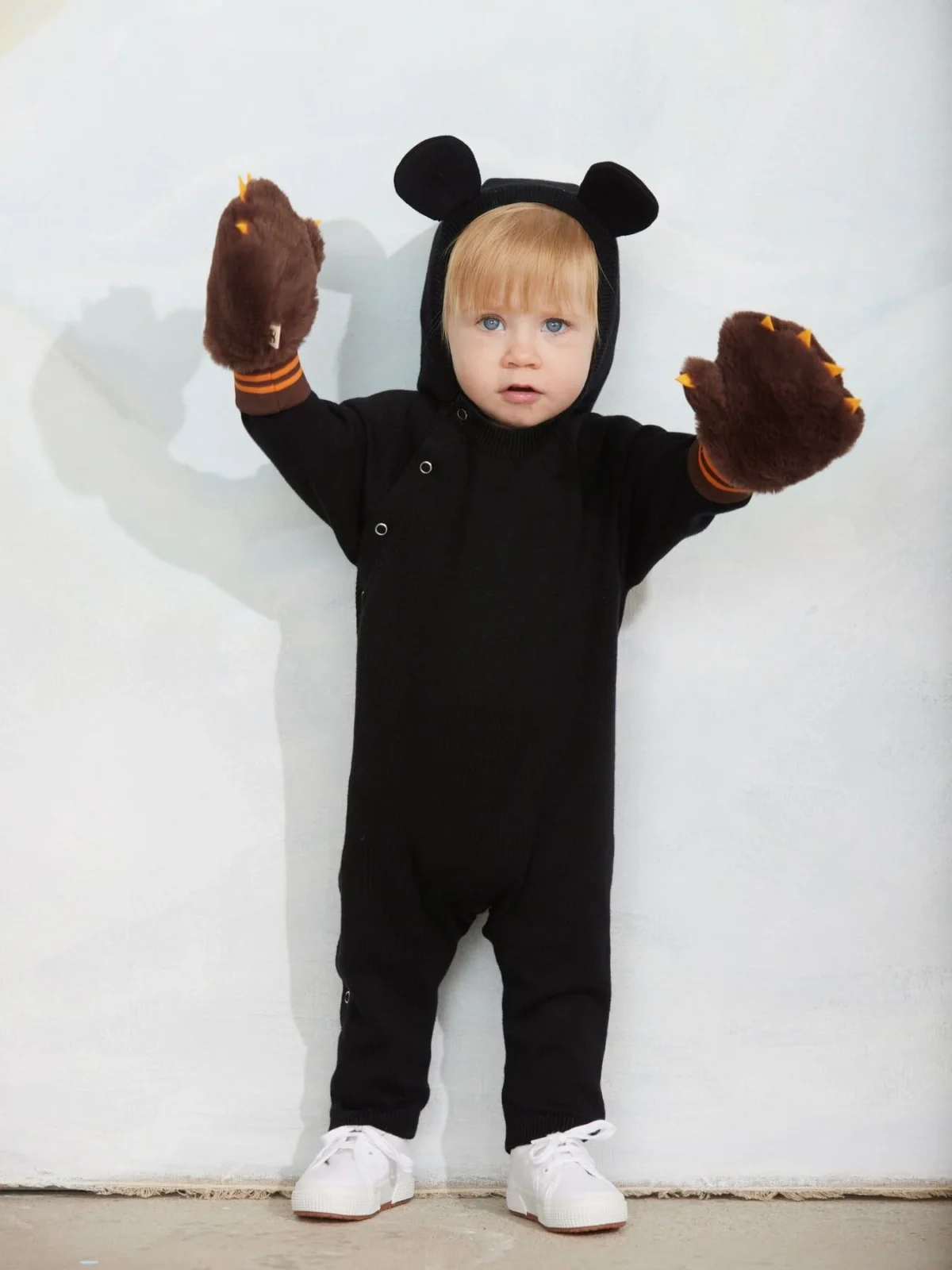 A toddler in a black hooded outfit with bear ears and brown paw gloves stands against a light background, arms raised, wearing white sneakers.