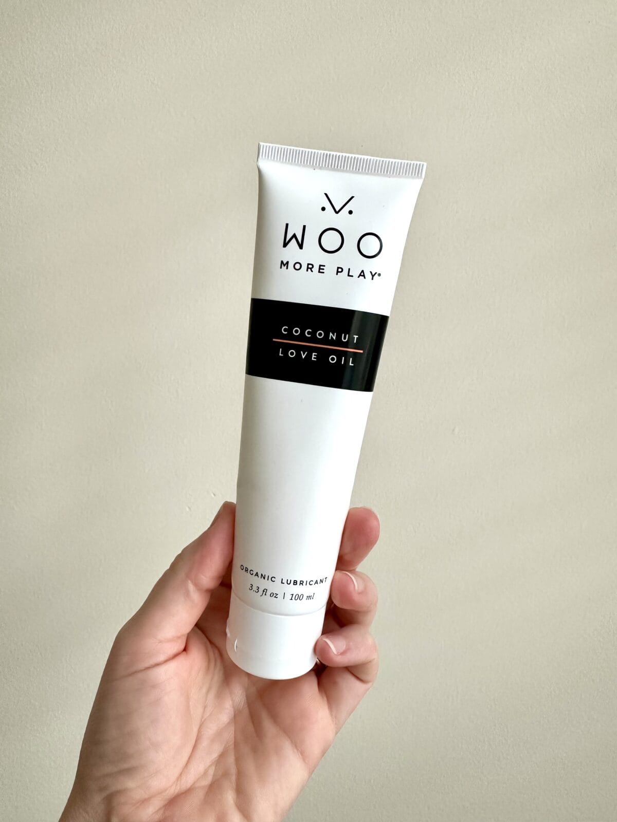 A person is holding a white squeeze tube of Woo More Play Coconut Love Oil organic lubricant against a cream-colored background.