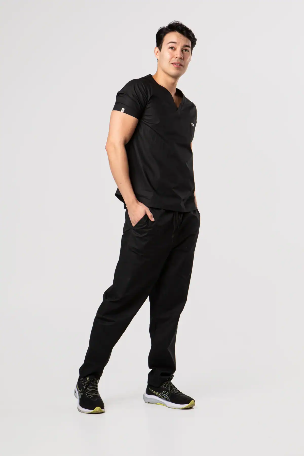 A smiling man with short dark hair is standing in a black medical scrub set from Percent Apparel with his hands in his pockets, posing against a plain white background.