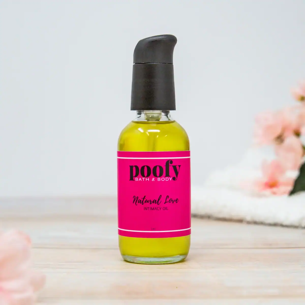 A clear glass bottle of bright yellow-green Poofy Bath & Body Natural Love Intimacy Oil with a bright pink label is featured on a light wooden surface with white and pink floral accents in the background.