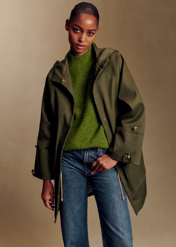 A person with a neutral expression is wearing a bright green sweater and an olive green, hooded, non-toxic winter coat with drawstrings, over a pair of blue jeans.