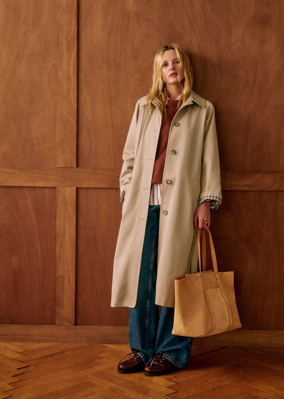 A person with long blonde hair and a neutral expression is standing against a wood-paneled wall and wearing a long, tan, non-toxic winter coat from Sézane, a brown sweater, a striped shirt, wide-leg blue jeans, and brown shoes, while holding a large, tan tote bag.