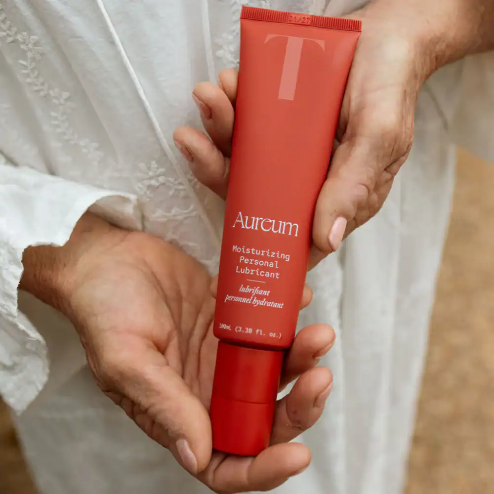 A mature hand is gently holding a coral-red tube of Aureum Moisturizing Personal Lubricant against a background of white flowing fabric.