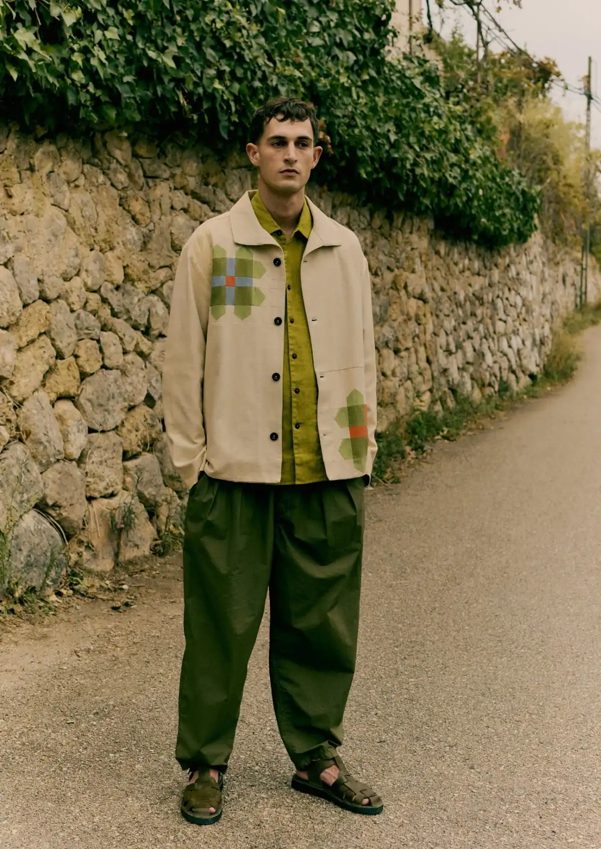 A man with a serious expression is standing on a narrow road in front of a stone wall covered in greenery, wearing a light-colored jacket with patches from Toast, a green shirt, green pants, and dark sandals.