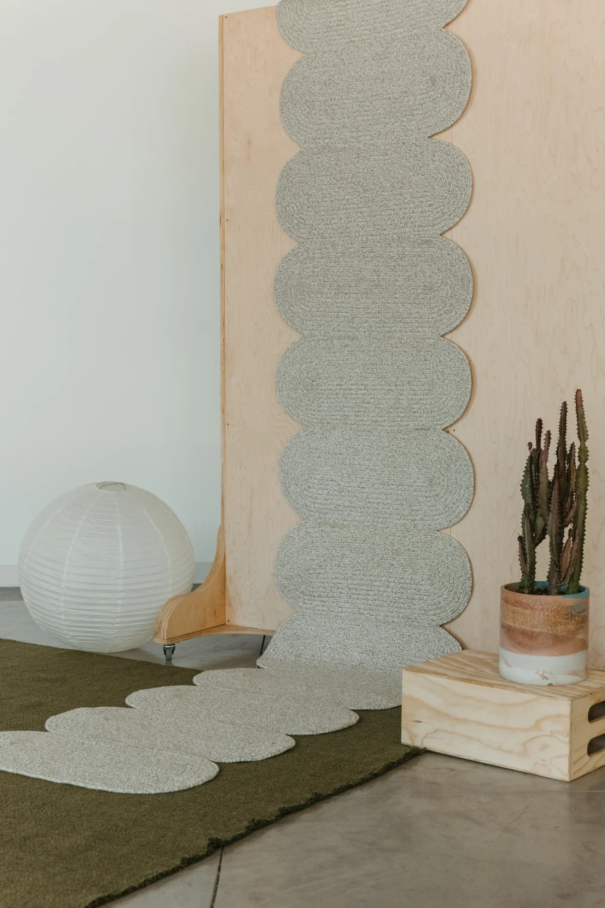 The photo features a large, light-colored paper lantern and a tall cactus in a terracotta pot on a wooden box, with a vertically cascading, scalloped light green runner rug set against a tall sheet of plywood, and a darker green rug covering the concrete floor in the foreground.