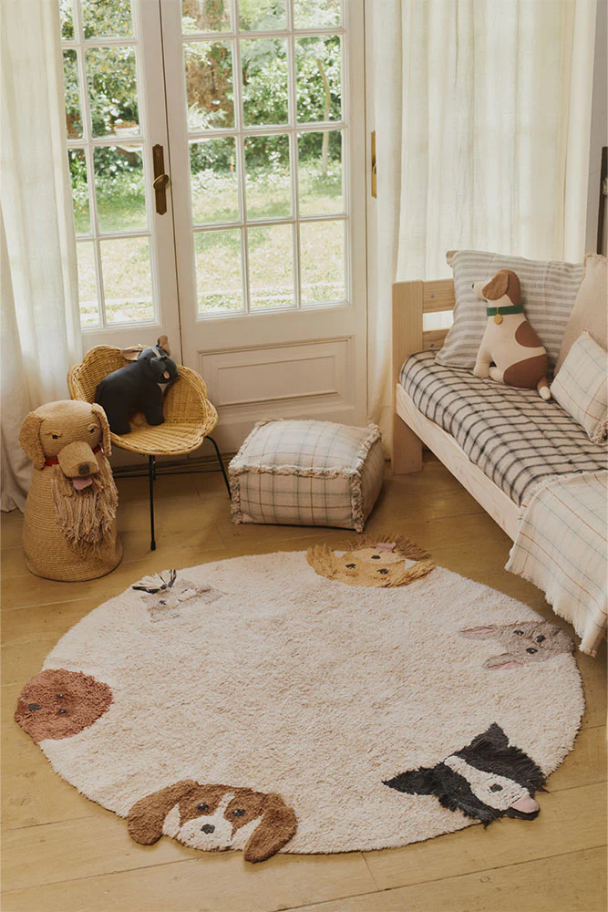 The photo captures a brightly lit child's room featuring a round rug decorated with dog and rabbit faces, a small bed with plaid bedding and dog plushies, a wicker chair holding a dark stuffed animal, and a glass-paneled door leading to an outdoor garden.
