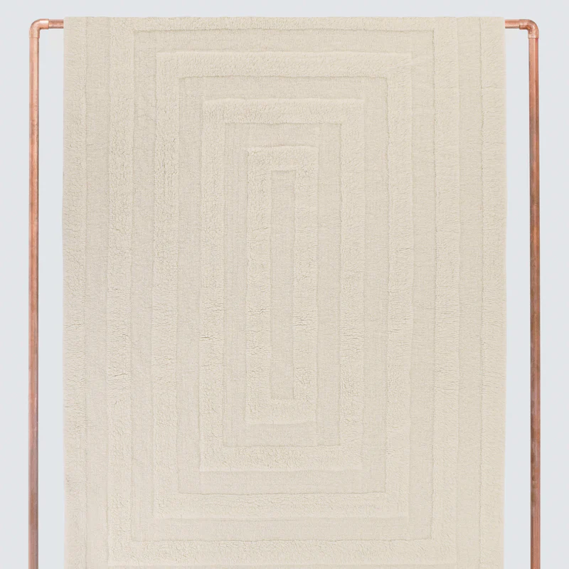 The photo displays a cream-colored, machine-washable rug with a subtle recessed rectangular pattern, hung vertically for presentation within a copper-colored metal frame