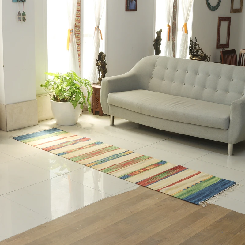 The photo shows a colorful runner rug with horizontal stripes in white, yellow, red, blue, and green, laid out on a white tiled floor in a bright room with a light gray sofa and a potted plant.