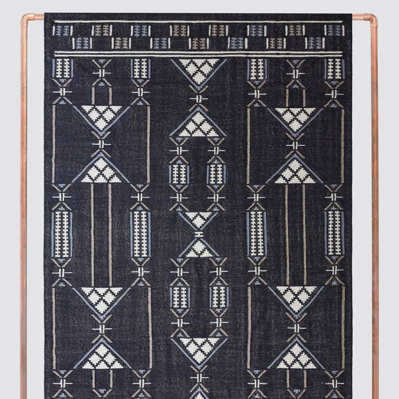 The photo shows a dark blue rug with white and light blue geometric, hourglass-like patterns, hung vertically from a copper-colored metal frame for display.
