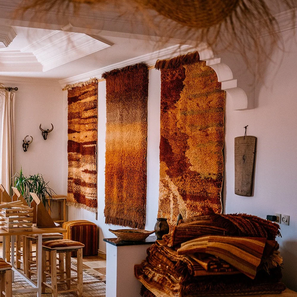The photo shows a room with a white arched doorway opening into a space decorated in warm, earthy tones, featuring three vertical, textural rugs hung on a white wall, a stack of rugs in the foreground, and carved wooden furniture with decorative animal skulls on the wall.