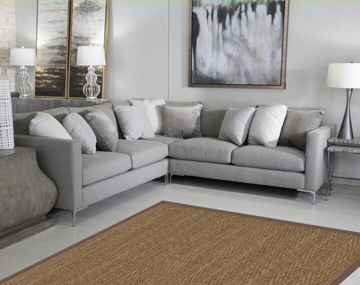 The photo displays a modern living room featuring a large, light gray sectional sofa, numerous decorative pillows in white and gray textures, abstract art on the wall, and a textured, woven brown area rug on a light tile floor.