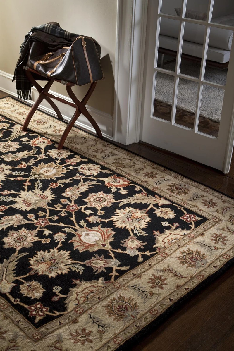 The photo captures a view through a doorway into a foyer or hallway, featuring a dark, traditional floral area rug with a wide, tan border placed on a dark wood floor, next to a white french door and a luggage rack with a bag and scarf.