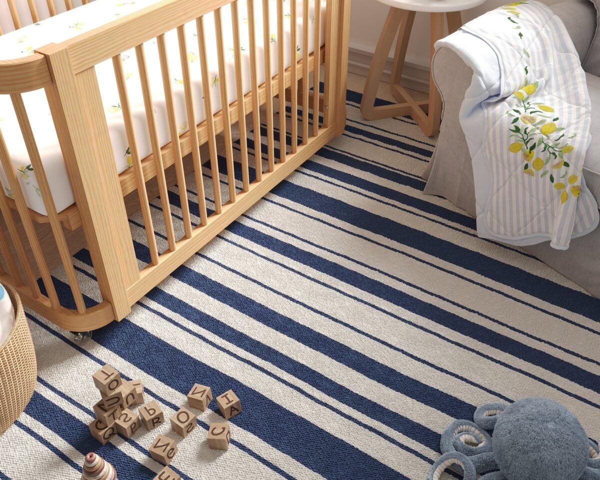 The photo captures a close-up of a natural wood crib with open slats and a striped rug in alternating white and navy blue stripes on the floor below, with a pile of wooden alphabet blocks visible on the rug.