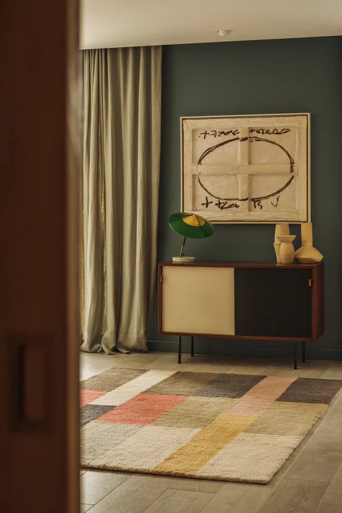 The photo captures a view through a doorway into a mid-century modern-styled room featuring a colorful, geometric rug, a dark green wall, a wooden credenza topped with a sculptural lamp and yellow vases, and a large abstract painting.