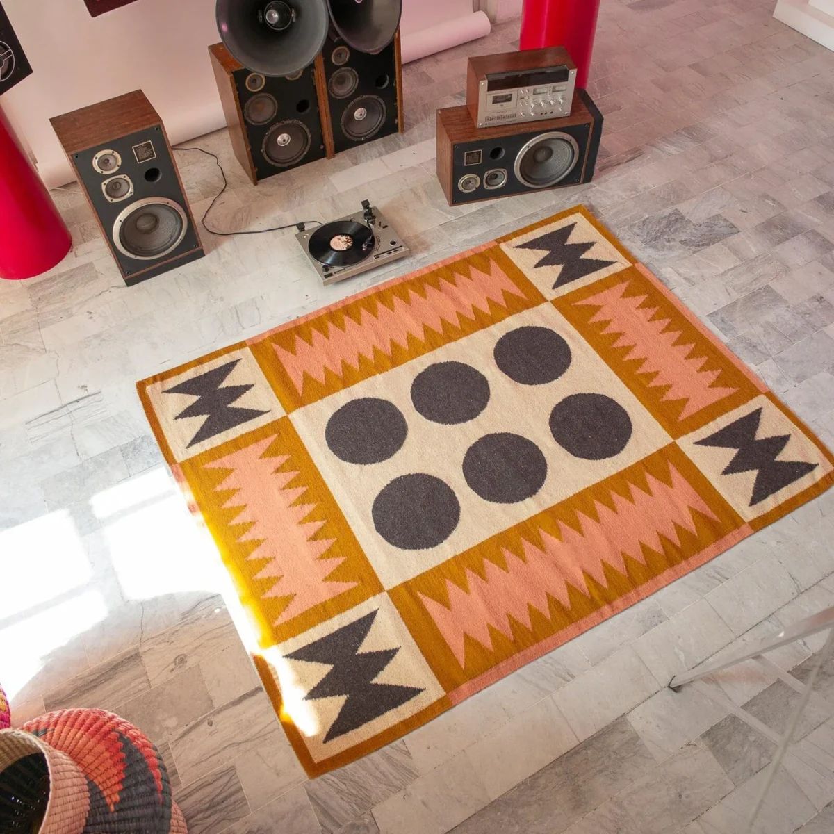 The photo displays a colorful, geometric rug with a pattern of dark circles and zigzags in shades of pink, tan, and dark gray, set on a tiled floor in a room with vintage-style audio equipment including speakers and a turntable.