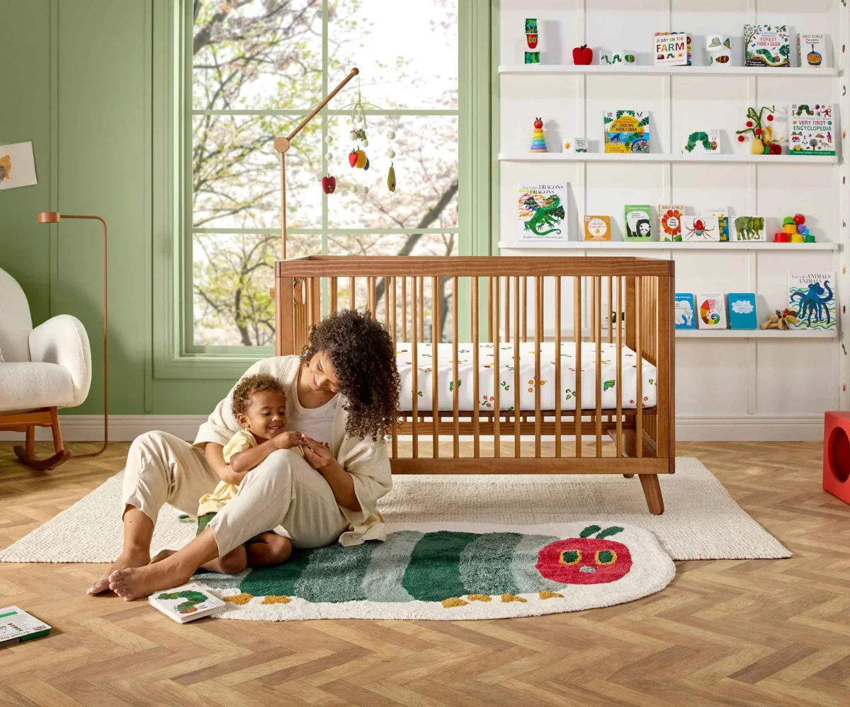 The photo shows a young woman sitting on a white rug, holding a smiling baby on her lap, next to a wooden crib and an Eric Carle's The Very Hungry Caterpillar themed rug in a bright room with shelves of children's books.