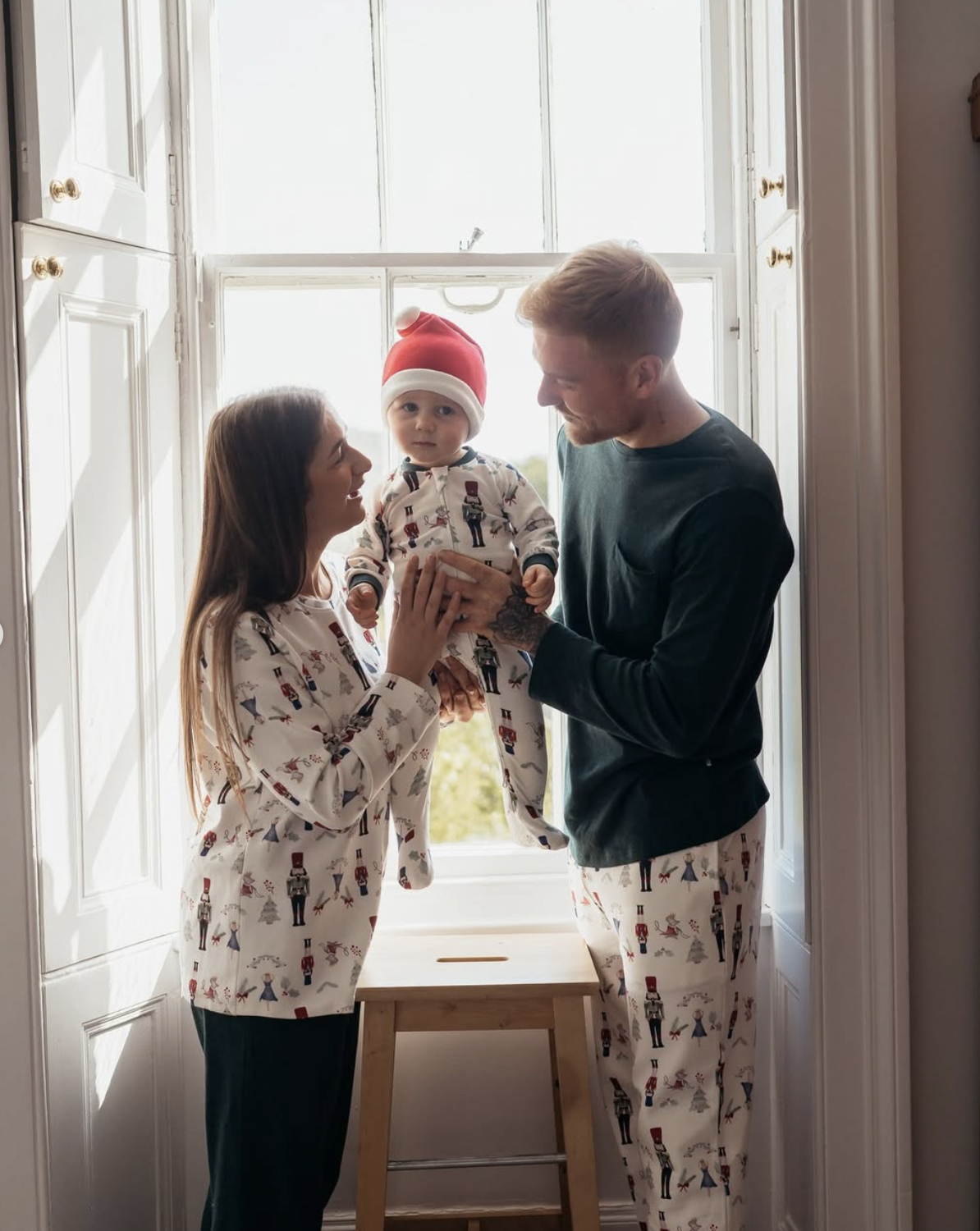 A family of three—a mother, father, and a baby in a Santa hat—are standing by a bright window, all wearing nutcracker-patterned pajamas, with the father lifting the baby above a wooden stool.