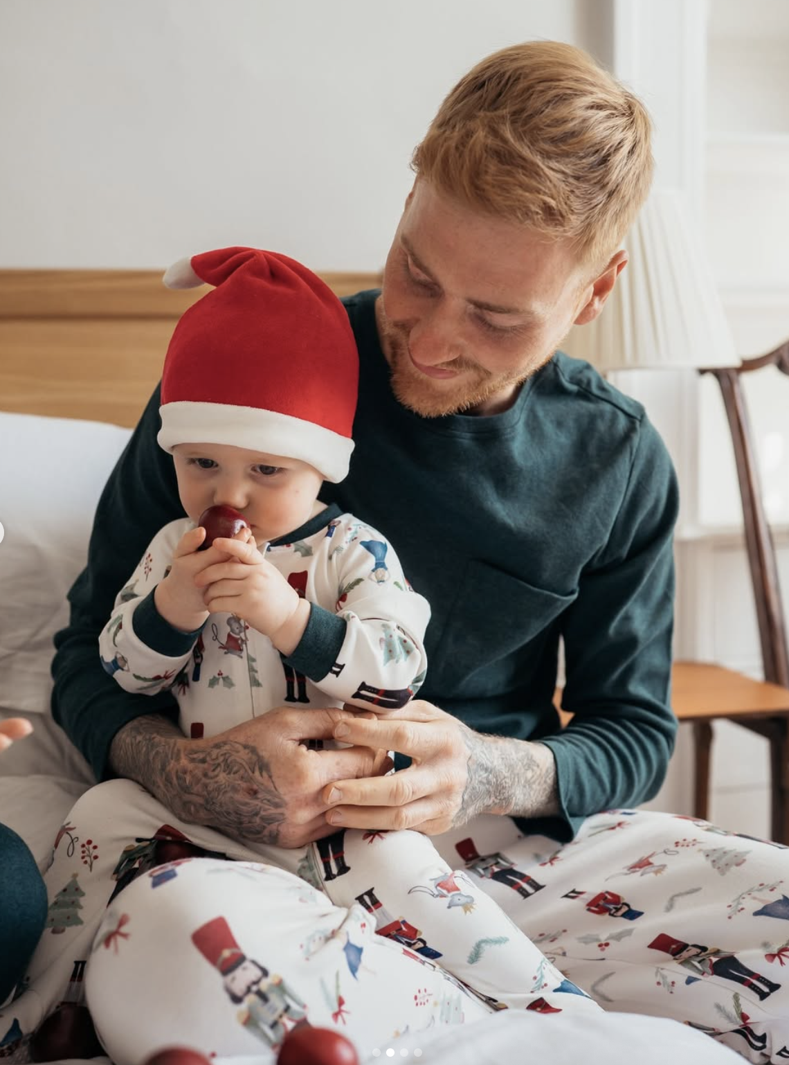A father with tattoos and a green shirt is smiling down at a baby in a Santa hat and nutcracker-themed pajamas who is seated on his lap and playing with a round red object.