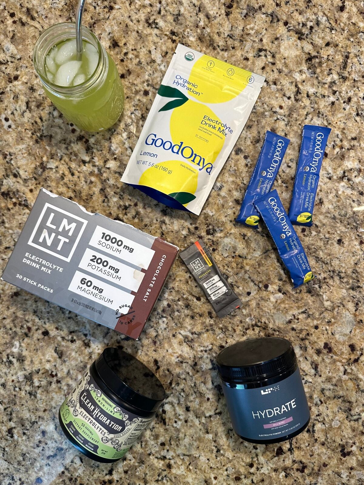 An overhead shot displays a collection of various electrolyte drink products, including GoodOnya Lemon mix, LMNT Chocolate Salt stick packs, LTH Hydrate, and another dark jar on a speckled countertop, next to a glass of what appears to be a green-hued prepared drink.