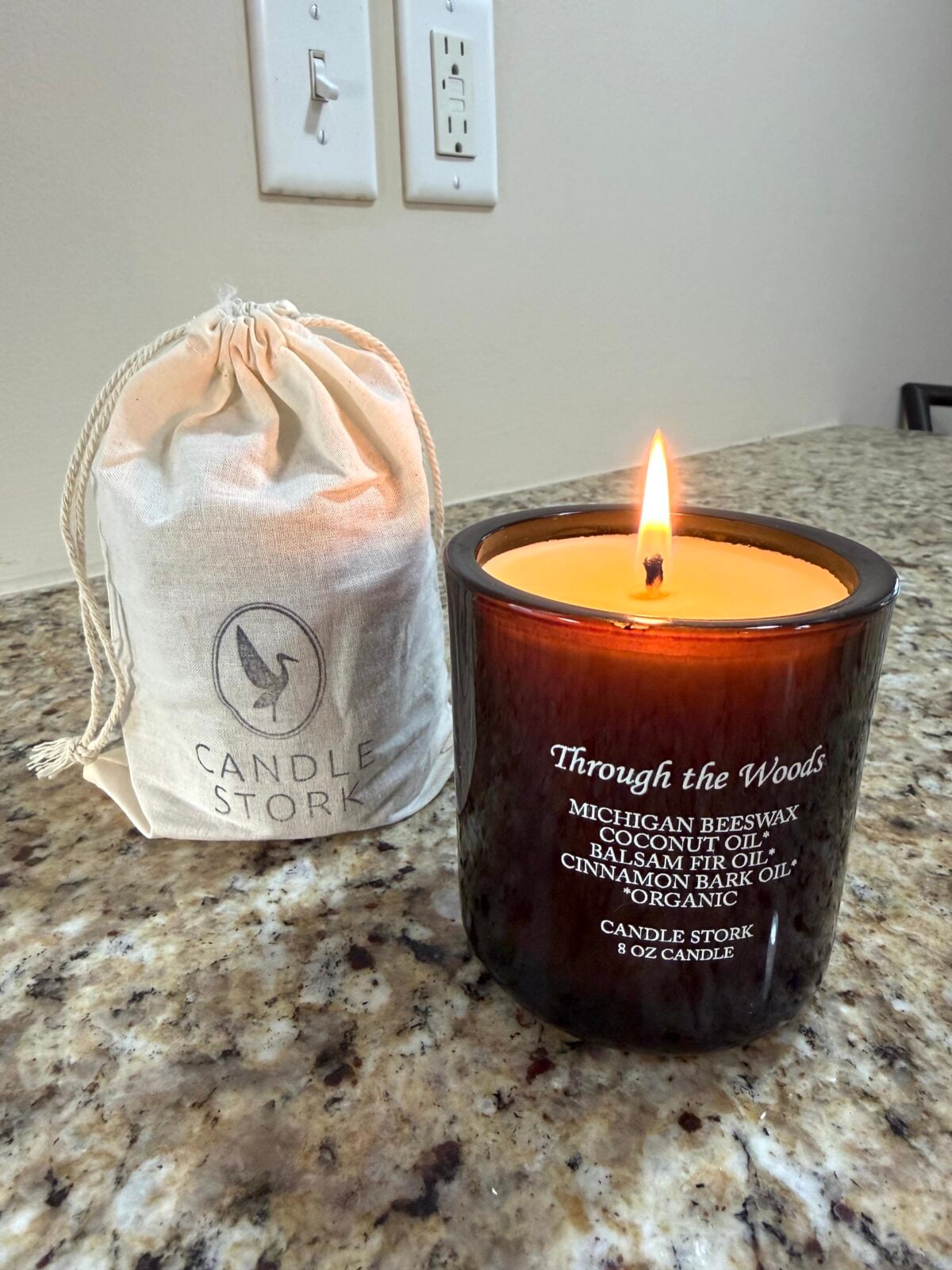 Lit beeswax candle in an amber glass jar labeled &ldquo;Through the Woods&rdquo; by Candle Stork, placed on a granite countertop beside a small drawstring canvas bag with the Candle Stork logo.