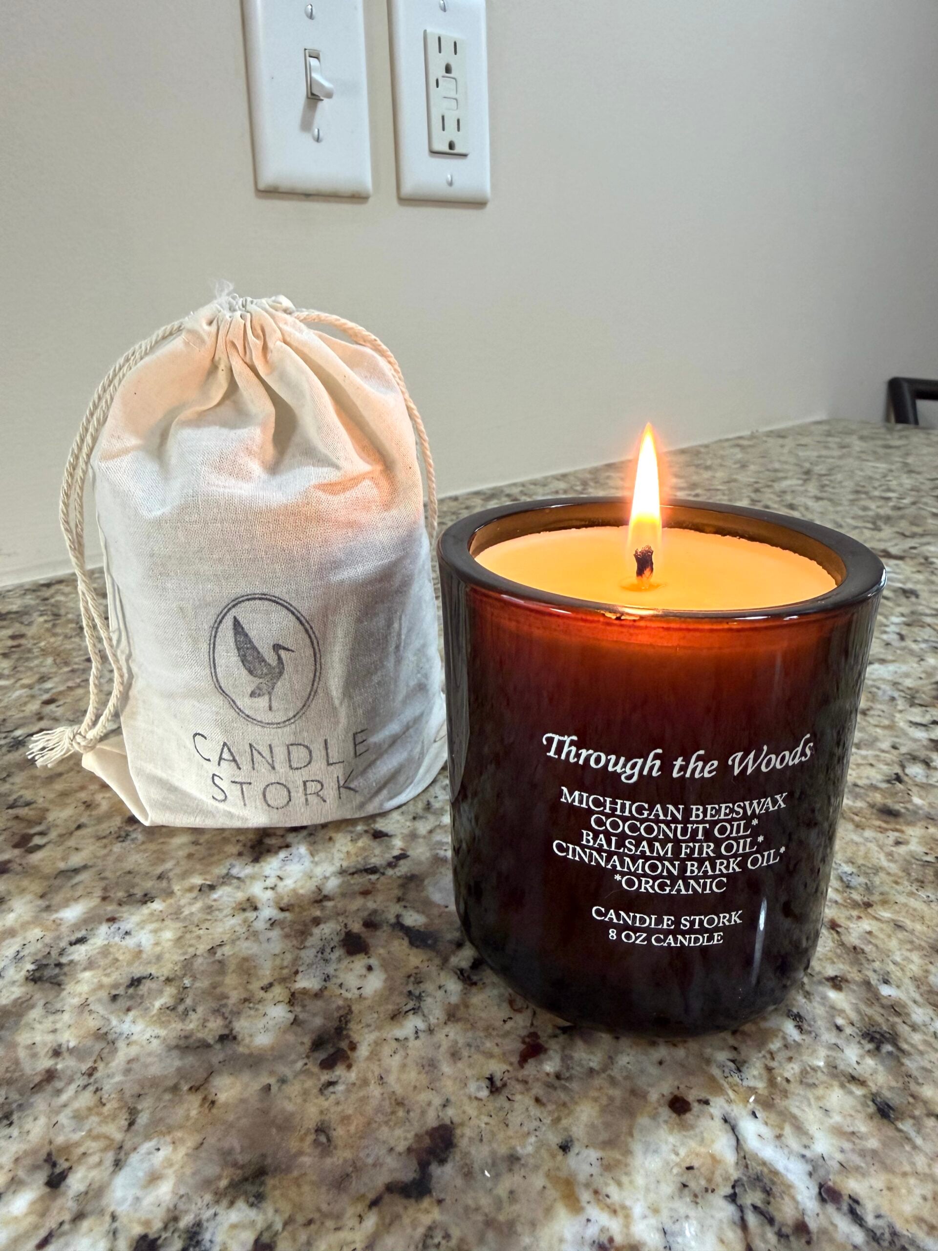 Lit beeswax candle in an amber glass jar labeled &ldquo;Through the Woods&rdquo; by Candle Stork, placed on a granite countertop beside a small drawstring canvas bag with the Candle Stork logo.