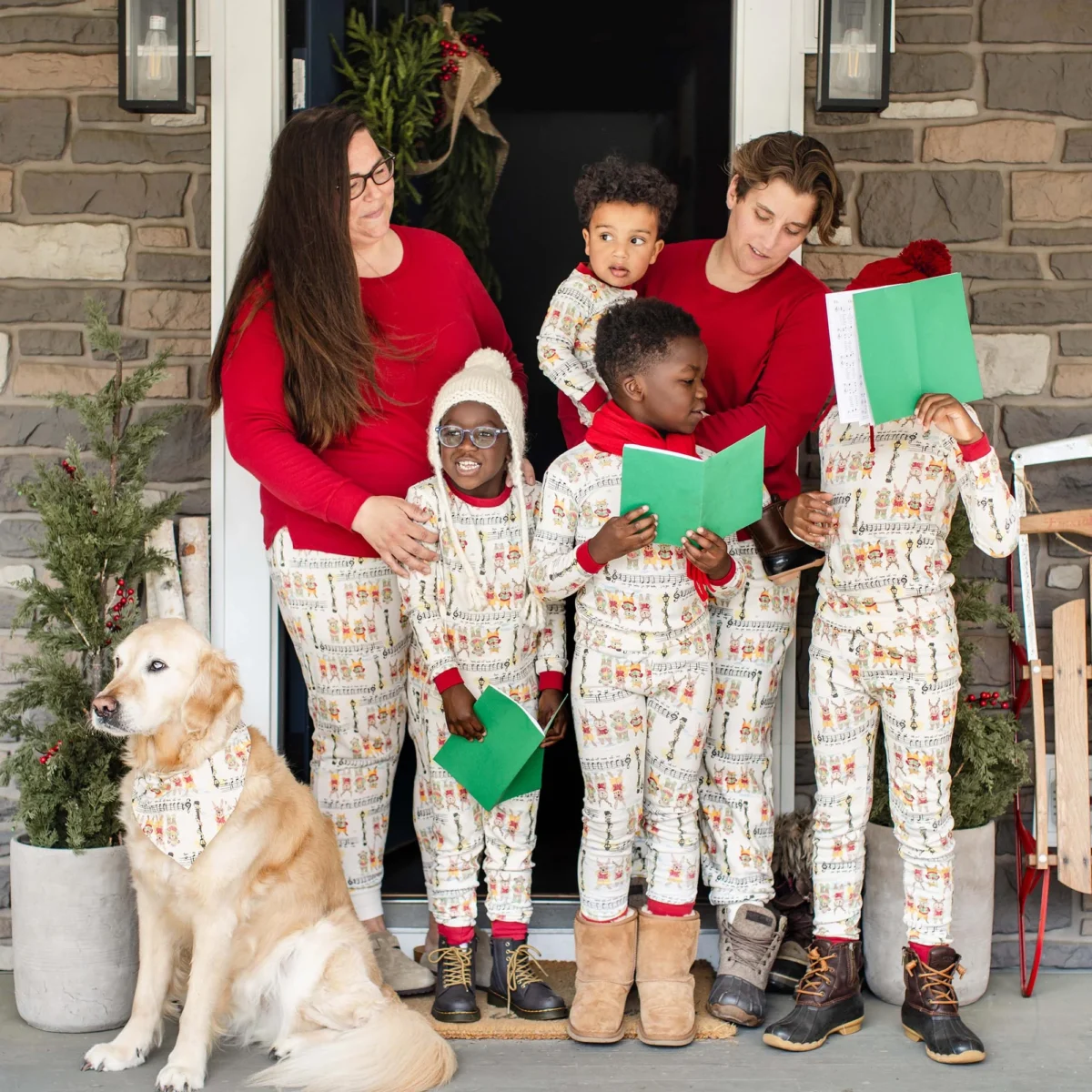 A family of six—two adults and four children, along with a golden retriever—is posing on a front porch, with the adults in red long-sleeved shirts and the children in matching patterned pajamas, holding open hymnal-style books.