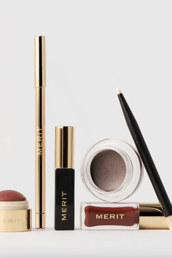 A collection of MERIT beauty products, including a cream blush stick, mascara, eyeliner pencil, brow gel, and a pot of cream eyeshadow with an applicator brush, arranged neatly on a light background.
