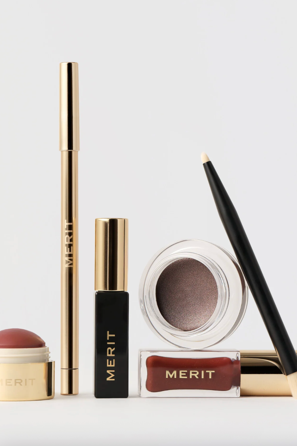 A collection of MERIT beauty products, including a cream blush stick, mascara, eyeliner pencil, brow gel, and a pot of cream eyeshadow with an applicator brush, arranged neatly on a light background.