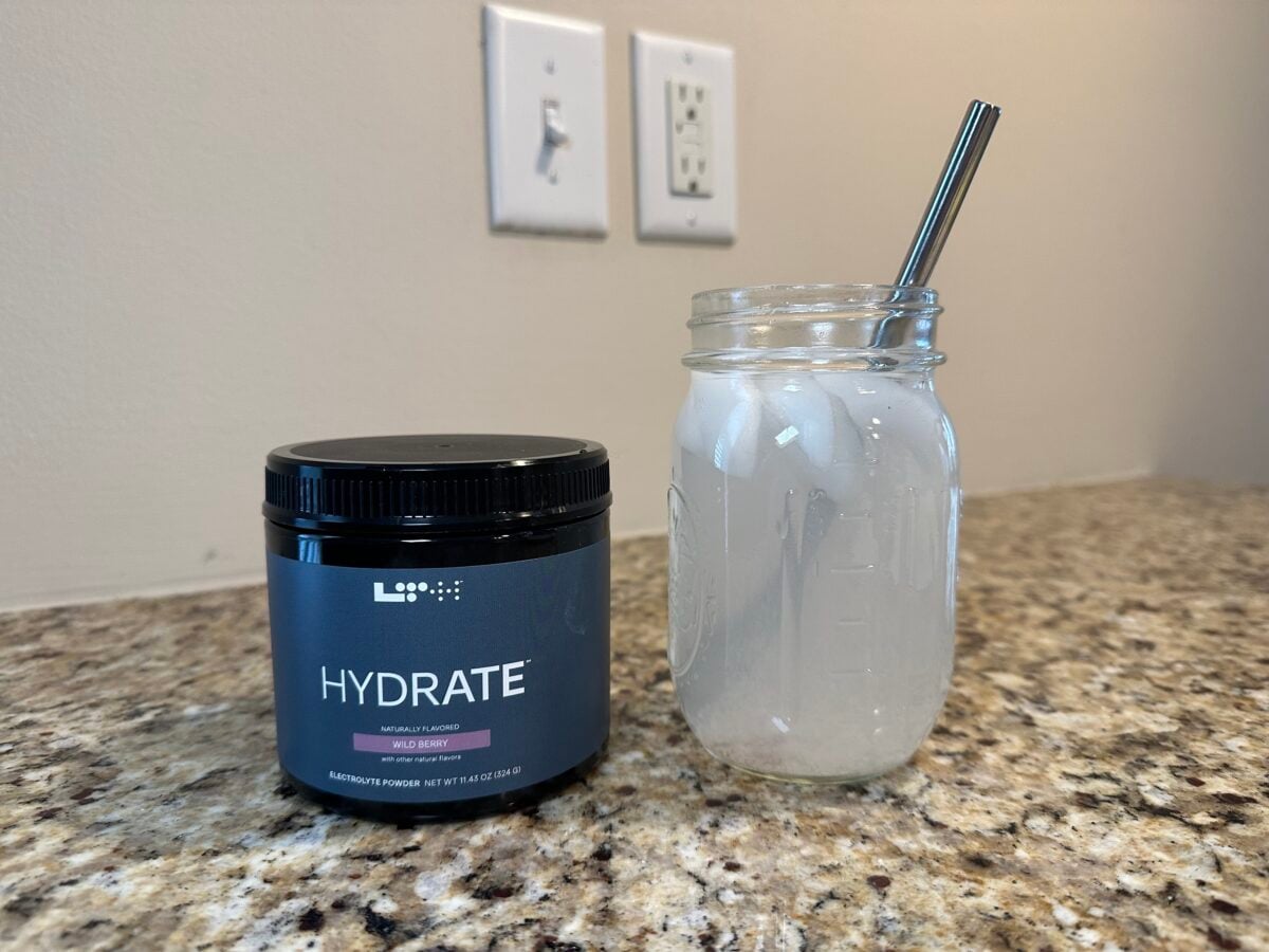 A dark blue jar of LTH Hydrate Wild Berry Electrolyte Powder sits on a speckled counter next to a glass of the prepared drink with ice and a metal straw.