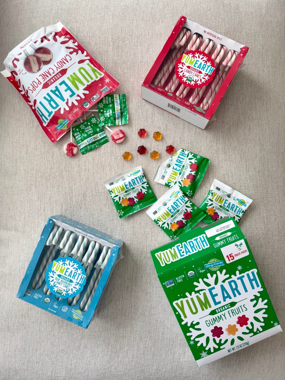 Assorted YumEarth holiday candies, including candy canes, candy cane pops, and gummy fruit snack packs, arranged on a light fabric background.