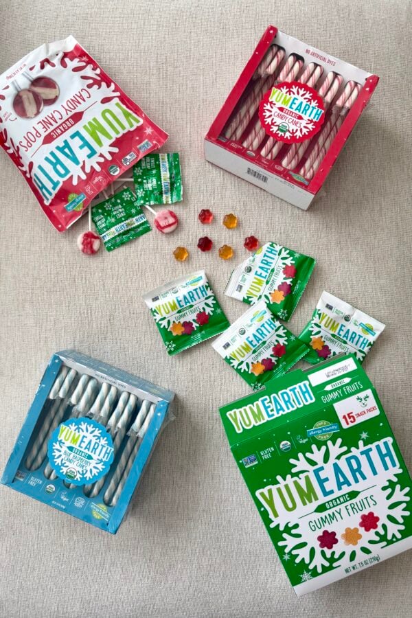 Assorted YumEarth holiday candies, including candy canes, candy cane pops, and gummy fruit snack packs, arranged on a light fabric background.