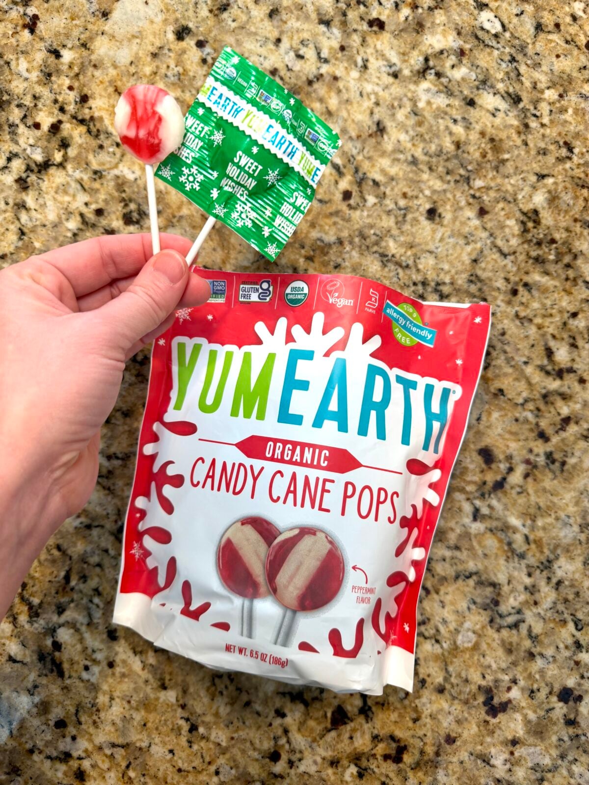 Hand holding a YumEarth organic candy cane pop and its green wrapper above a bag of YumEarth Organic Candy Cane Pops on a granite countertop.