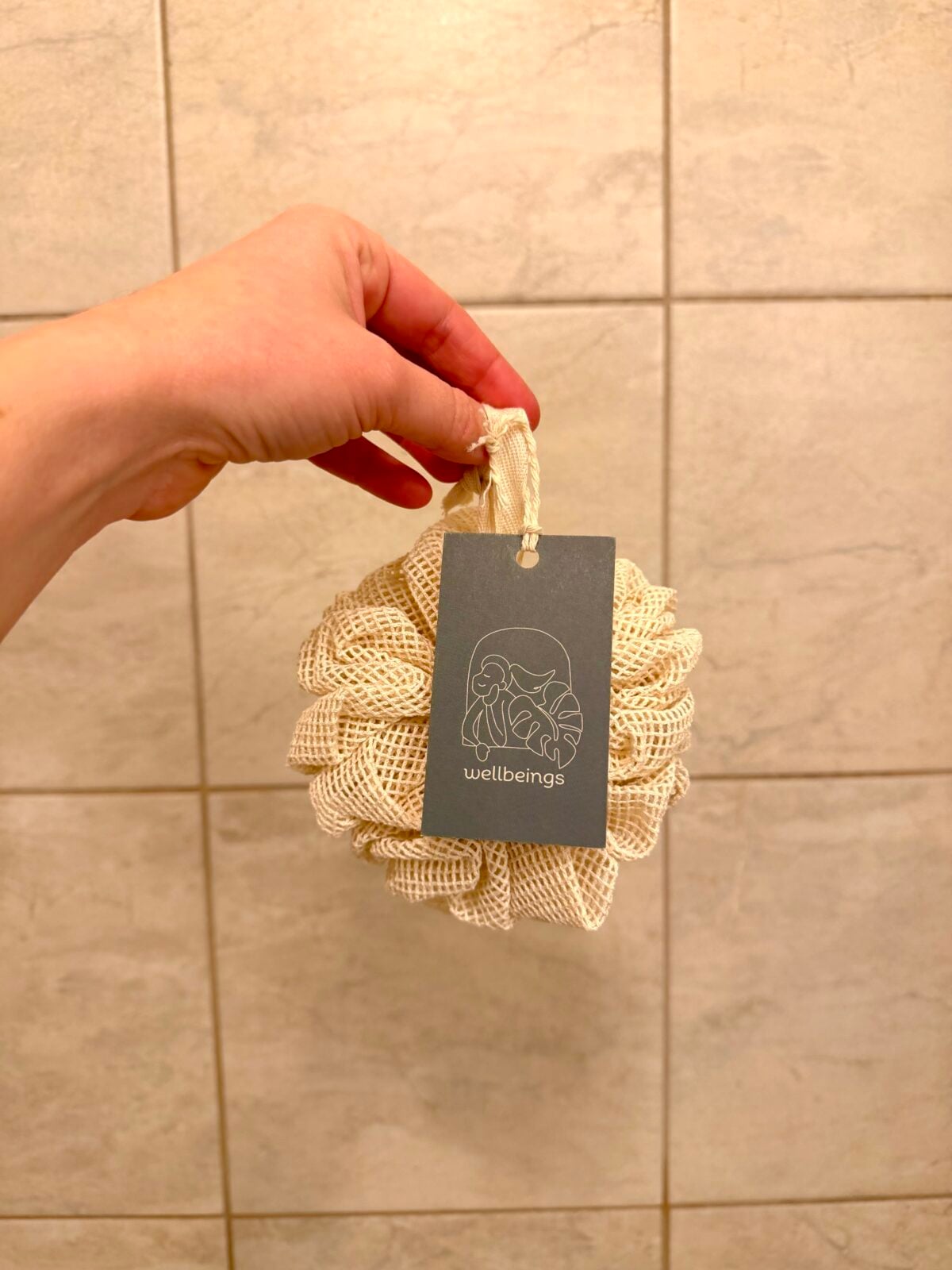 Hand holding a natural mesh loofah with a “wellbeings” tag against a tiled bathroom wall.