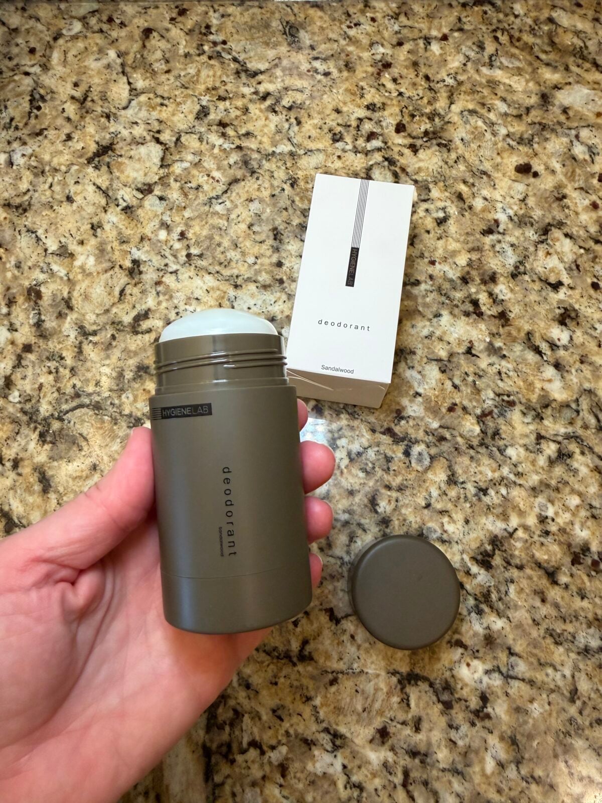 A hand holds an open container of sandalwood-scented deodorant by the brand Hygiene Lab, positioned next to its white box and cap on a speckled granite surface.