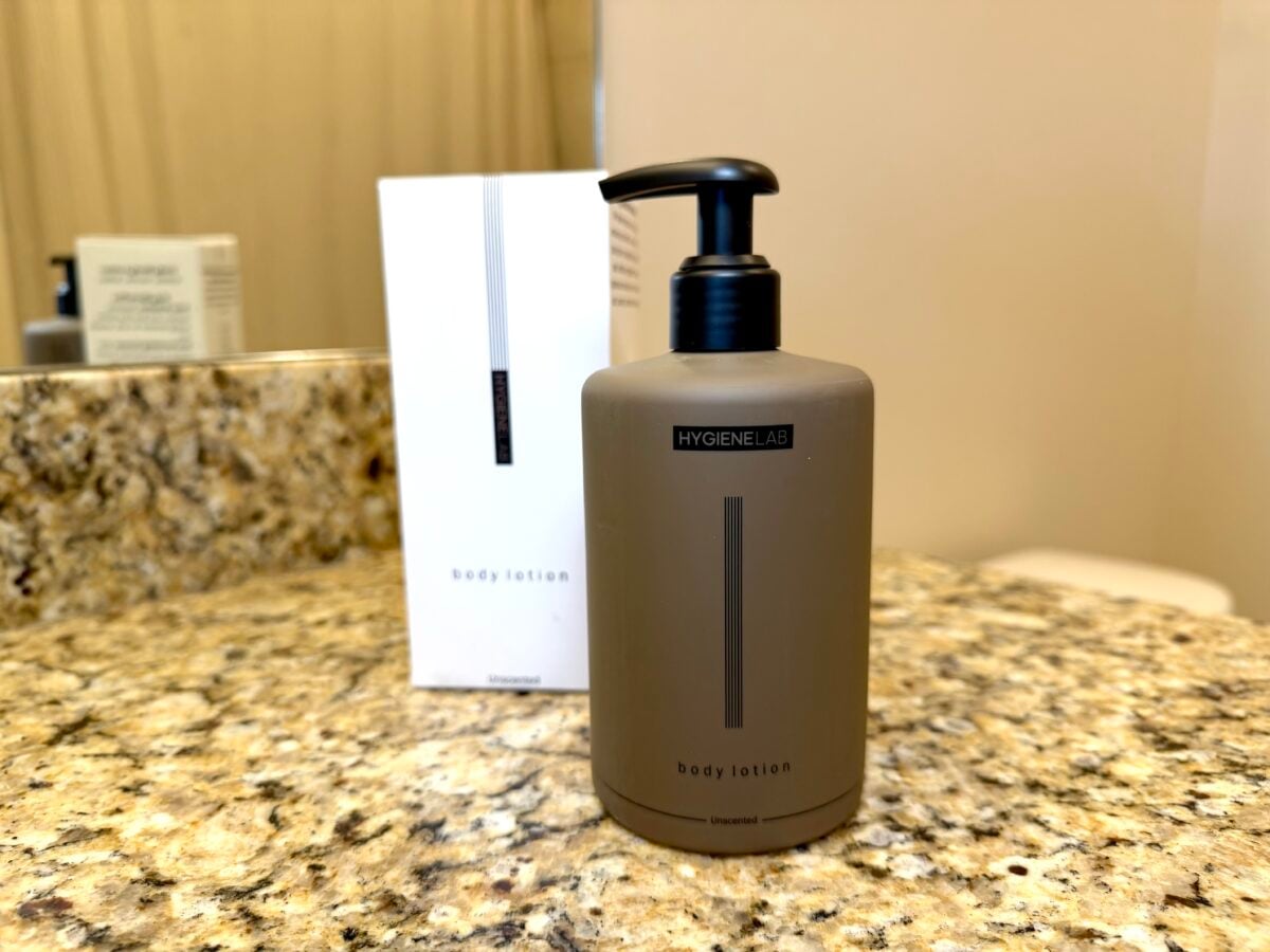 The photo features a matte brown pump bottle and its white cardboard packaging for Hygiene Lab body lotion sitting on a speckled granite bathroom countertop.